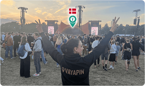 A woman in a VayaPin hoodie faces away from the camera, raising her arms in peace signs toward a large outdoor festival stage in Denmark.