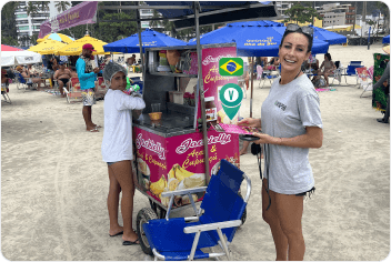 A smiling woman in a VayaPin t-shirt stands by a colorful food cart on a crowded beach in Brazil