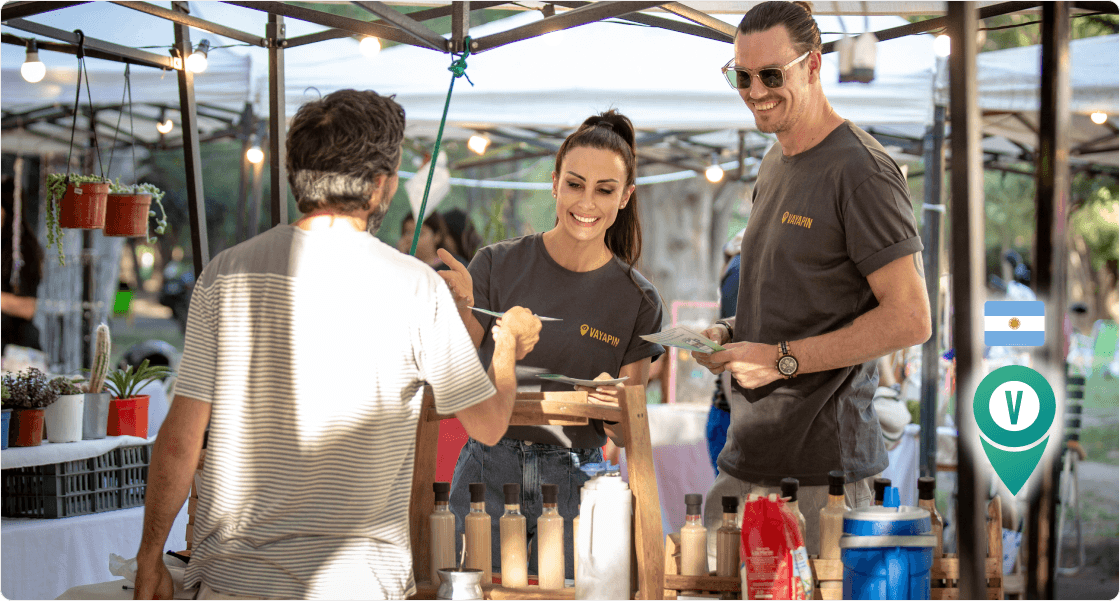 VayaPin founders smile while handing out flyers to a customer at an outdoor market stall in Argentina.