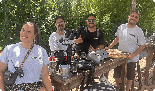A team of four VayaPin staff members, including a videographer with a gimbal, pose together at an outdoor wooden table