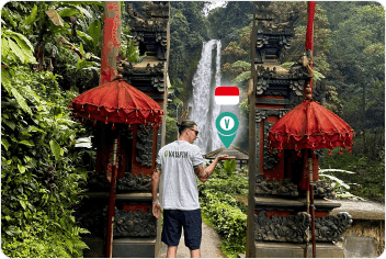 A man wearing a VayaPin t-shirt stands between ornate stone pillars overlooking a lush waterfall in Indonesia.