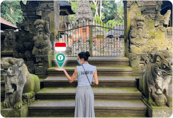 A woman in a VayaPin shirt stands before the ornate stone entrance and statues of a traditional Balinese temple in Indonesia