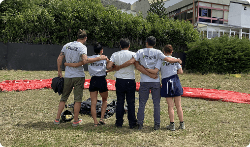A group of five people in VayaPin t-shirts stand with their arms around each other on a grassy field next to a red paragliding wing
